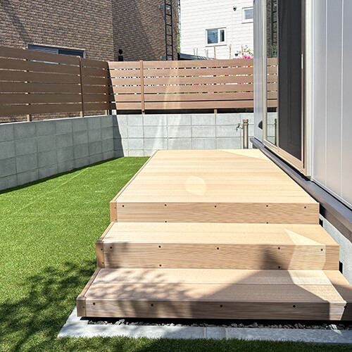 Wooden exterior steps leading to a raised deck beside a house with sliding glass doors, lawn on the left and a privacy fence in the background.