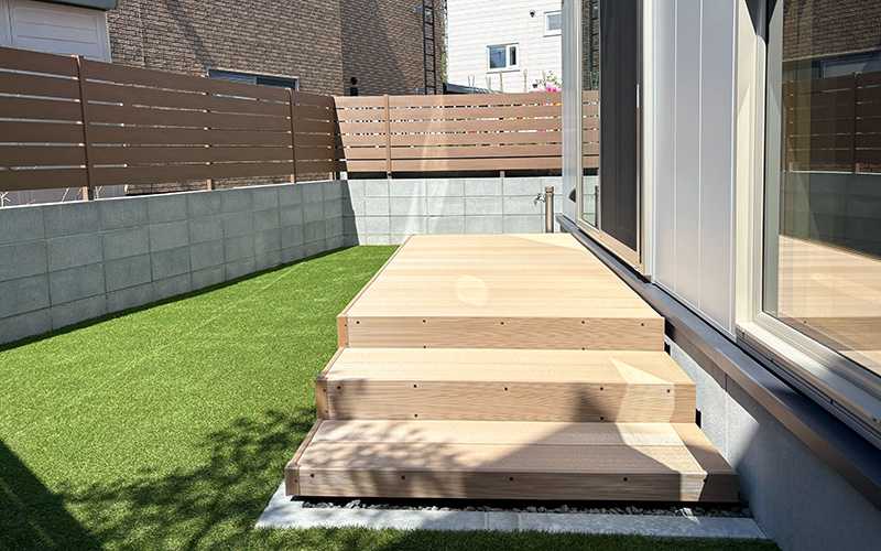 Wooden exterior steps leading to a raised deck beside a house with sliding glass doors, lawn on the left and a privacy fence in the background.