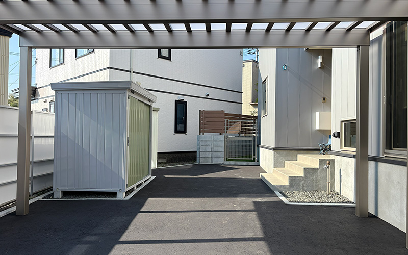 Paved driveway between modern white houses with a small gray storage shed on the left and concrete steps with a metal pergola overhead on the right.