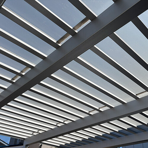 Metal pergola with parallel beams under a clear blue sky, creating a grid pattern of shadows and light on the structure.