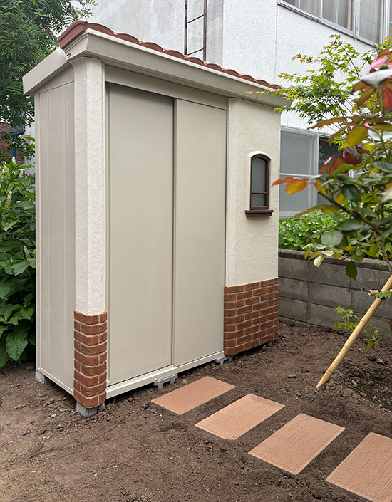 Beige outdoor storage shed with brick base and sliding doors in a garden, featuring a small arched window and a tile roof, with stepping-stone path nearby.
