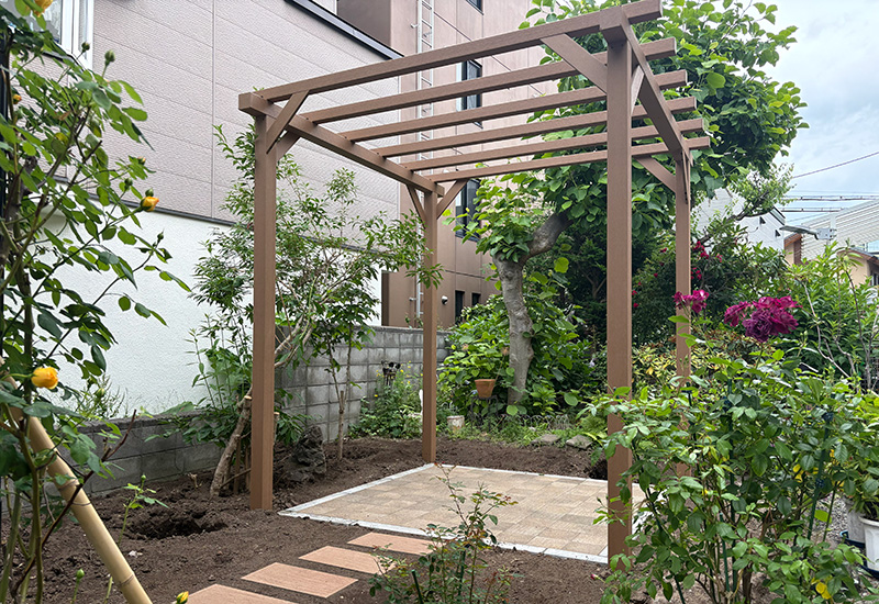 Wooden garden pergola over a beige tiled patio, surrounded by trees and flowering shrubs in a residential yard.