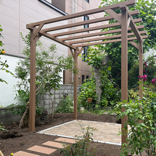 Brown wooden pergola over a small tiled patio in a garden, surrounded by trees and flowering shrubs.