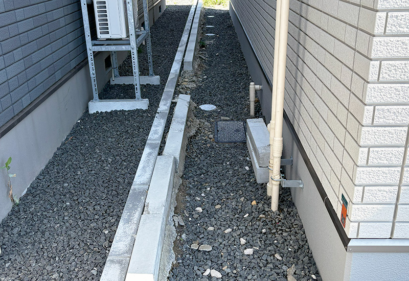 Narrow gravel alley between two walls with exposed pipes and an outdoor unit mounted on a rack.