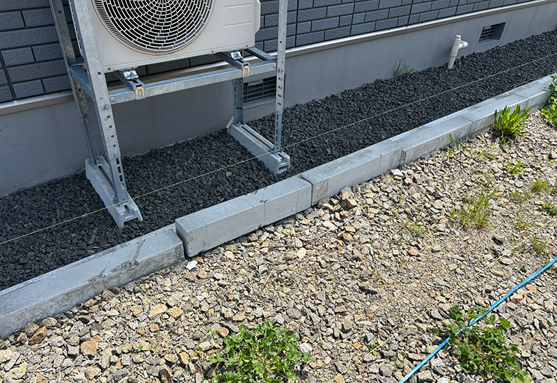 Outdoor air conditioning condenser on a metal frame beside a gravel yard, bordered by concrete blocks with a few plants nearby.