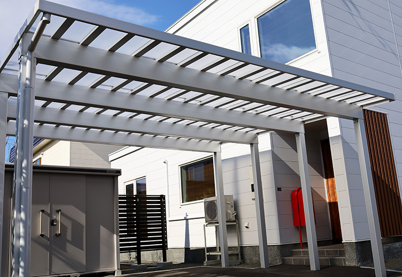Modern white house exterior with a gray metal pergola over a small patio, red mailbox by the entryway, and outdoor equipment nearby.