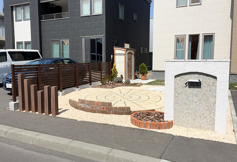 Residential front yard with a circular beige stone patio, brick edging, wooden slat fence, and a stone pillar mailbox.
