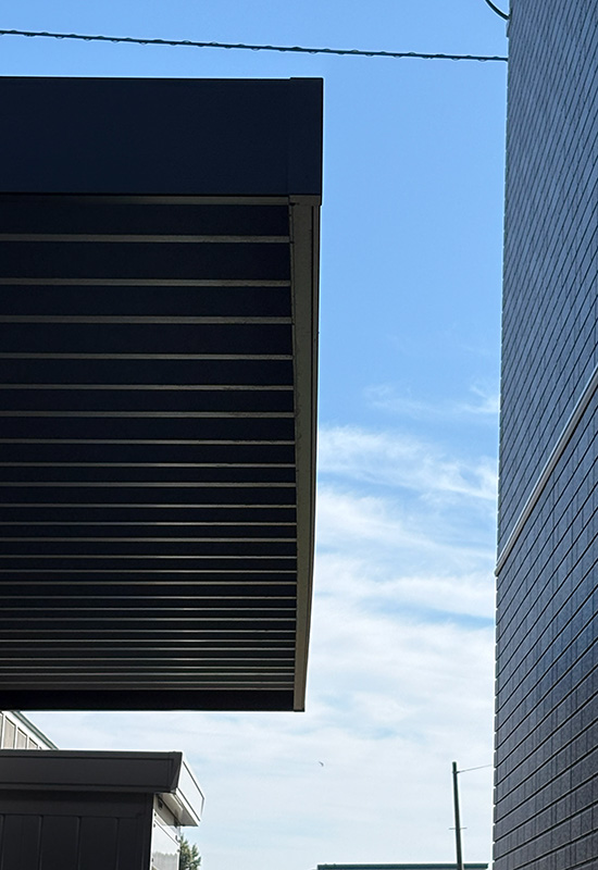 Close-up of a dark cantilevered building overhang with metal beams, against a blue sky and a brick wall to the right.