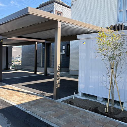 Carport with metal posts beside a modern brick building, a gray utility shed, and a small tree on a paved walkway area.