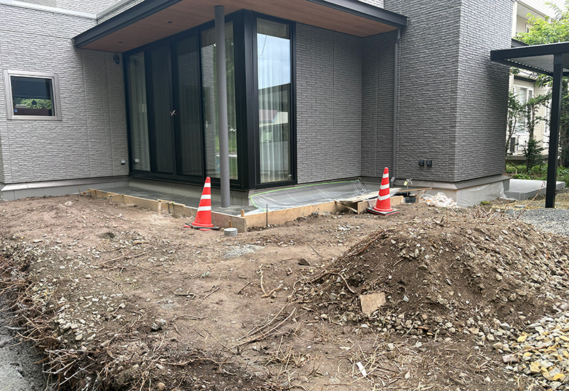 Back patio under construction: dirt yard, forms around a newly poured concrete slab, and two orange traffic cones near sliding glass doors of a modern house