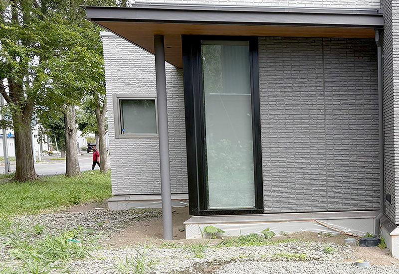 Corner of a modern gray brick house with a tall glass entry door under a canopy and a gray pillar nearby.