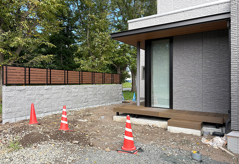 Modern house front with a wooden deck under construction, orange traffic cones on dirt yard, and a gray brick exterior.