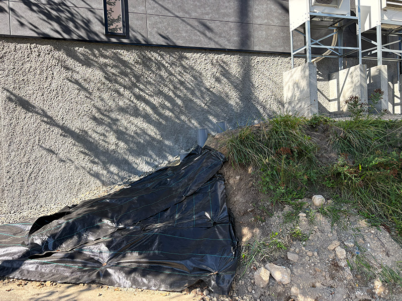 Black plastic tarp covering a dirt slope against a building wall, with scattered rocks and overgrown grass nearby near exposed scaffolding railings.