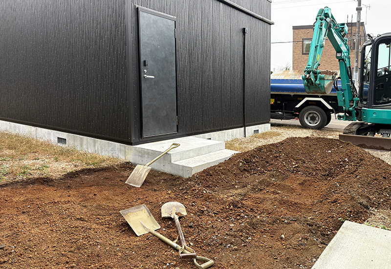 Construction site beside a black modular building; dirt pile, shovels, and a small excavator nearby.