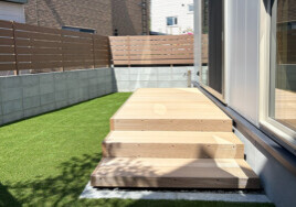 Wooden exterior steps leading to a raised deck beside a house with sliding glass doors, lawn on the left and a privacy fence in the background.